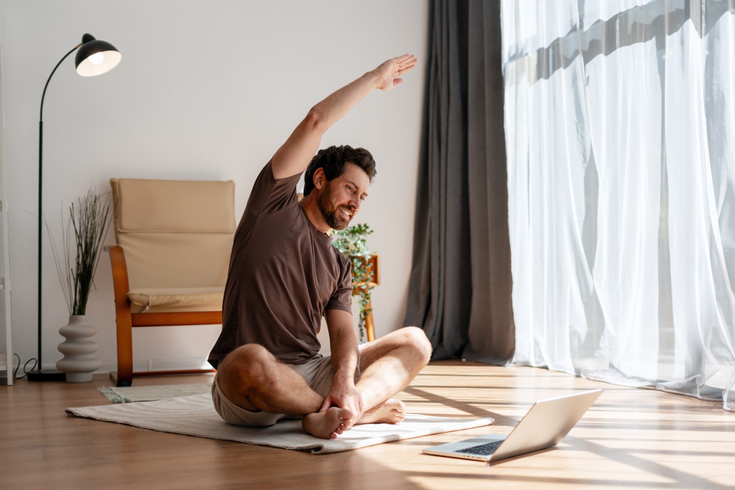 Man doing seated side stretch yoga pose on a mat near a laptop in a sunlit room with large window curtains.
