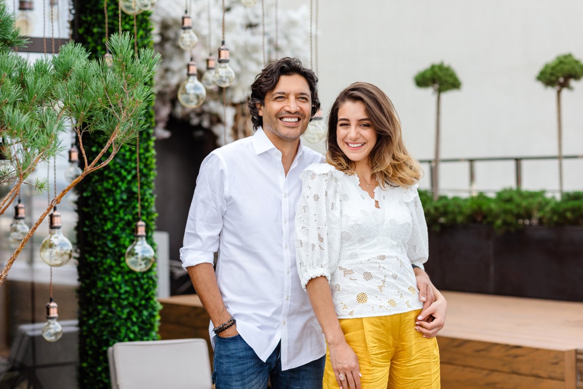 A smiling couple stands on a stylish patio with greenery and hanging lights. Both wear casual white tops; the woman wears yellow pants. Bright, joyful atmosphere.