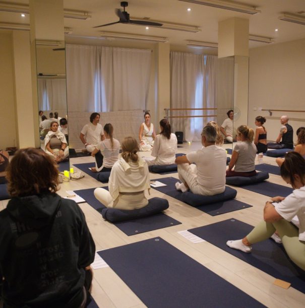 A serene yoga class with participants seated on blue mats in a dimly lit studio, surrounded by soft yellow candles. A calming and focused atmosphere.