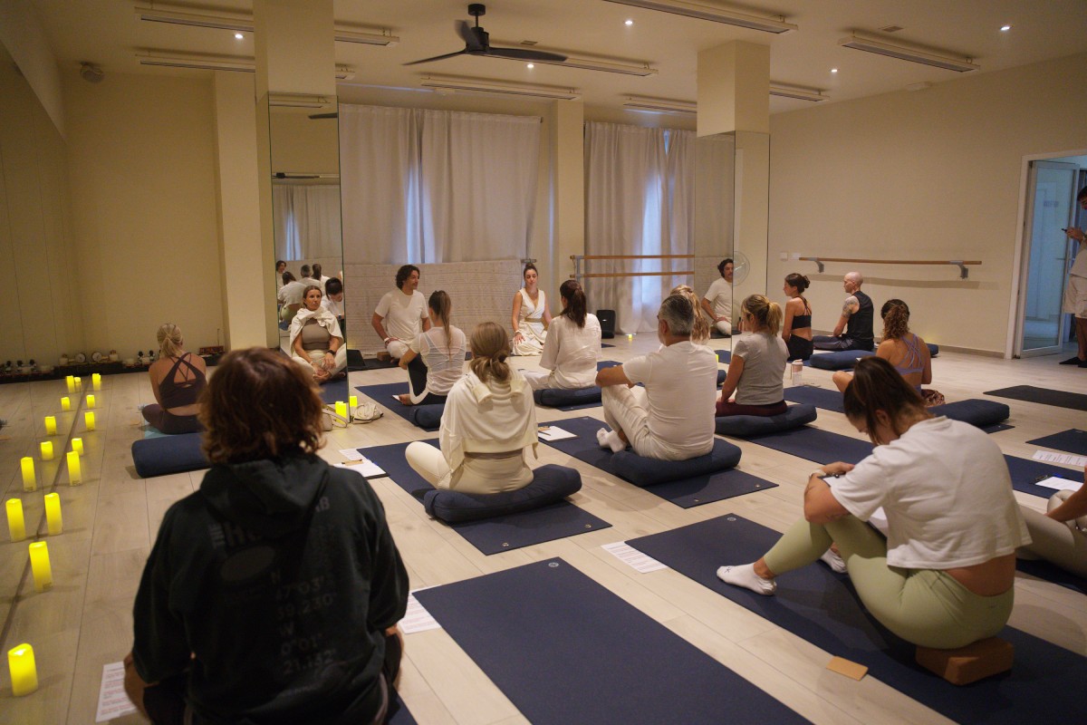 A serene yoga class with participants seated on blue mats in a dimly lit studio, surrounded by soft yellow candles. A calming and focused atmosphere.