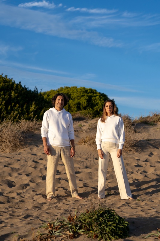 A man and a woman in white outfits stand on a sandy beach with sparse greenery. The sky is clear and blue, creating a serene and relaxed atmosphere.