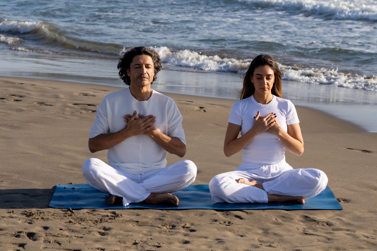 A man and woman sit cross-legged on a beach, meditating with hands on their chests, exuding calmness. The ocean waves gently roll in behind them.
