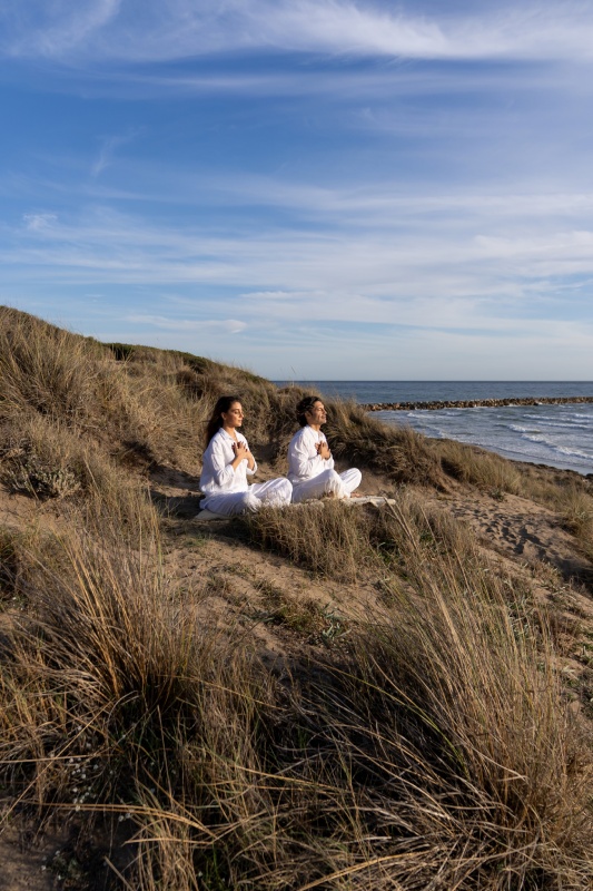 Two people in white attire meditate on grassy dunes by the sea under a clear blue sky, conveying a sense of calm and tranquility.