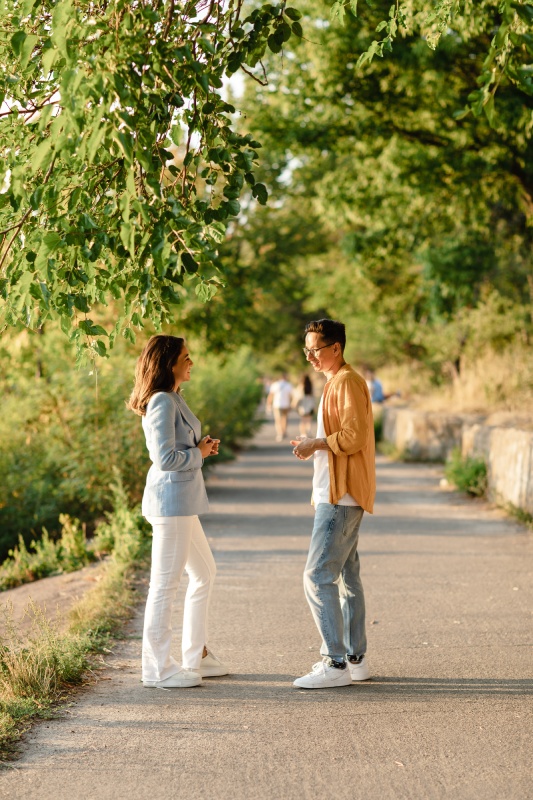 A man and woman casually chat on a sunlit path lined with green trees. They appear relaxed and engaged. Dappled sunlight creates a warm, pleasant ambiance.