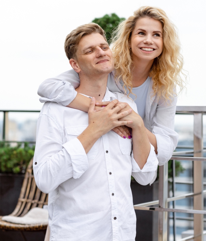 A smiling couple embraces on a rooftop. The woman hugs the man's shoulders from behind, both wearing white shirts. The mood is joyful and serene.