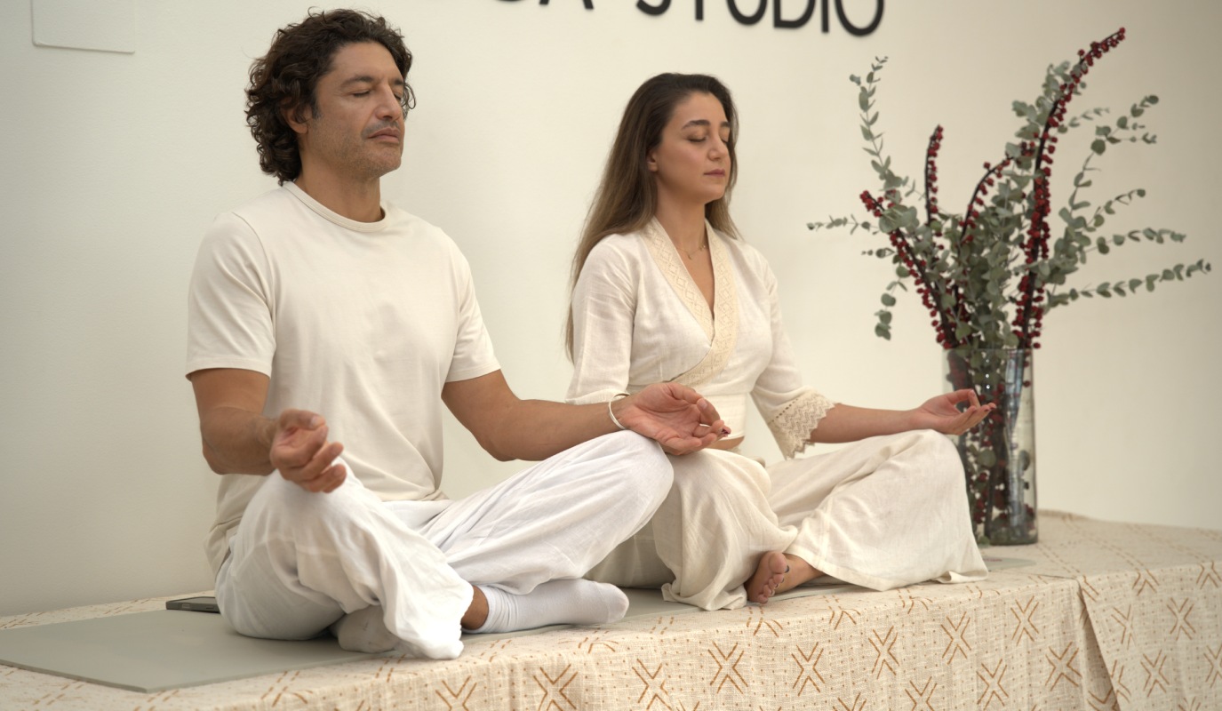A man and woman meditate in white clothing on a patterned mat, eyes closed, hands in mudras, evoking calmness in a serene yoga studio setting.