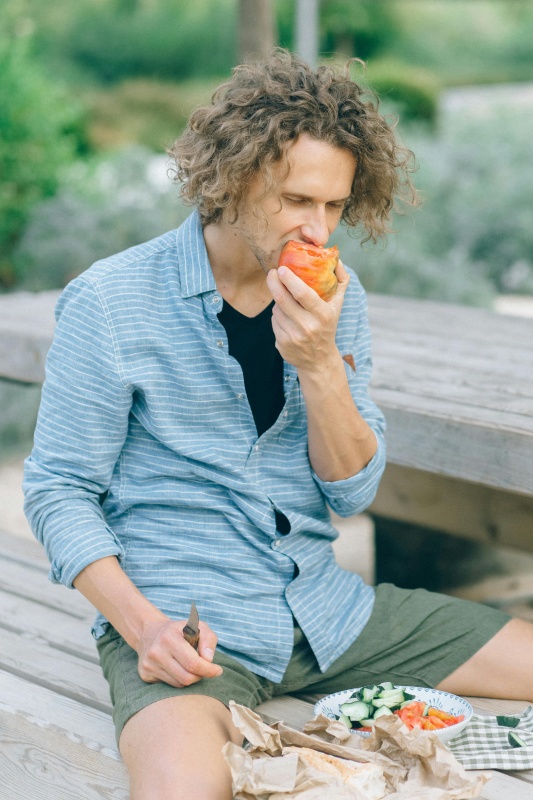 Person with curly hair sitting on a picnic bench eating an apple, with a plate of sliced vegetables nearby.