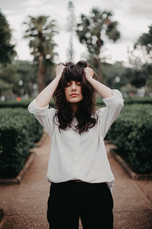 Woman with eyes closed holds head, expressing frustration or contemplation, standing on a garden path lined with bushes and palm trees.