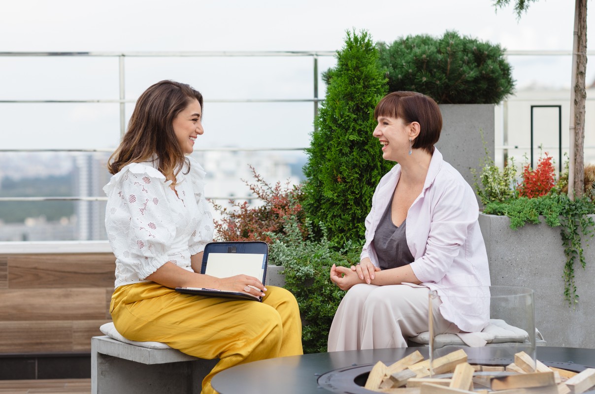 Two women sit outdoors, smiling and engaging in conversation. One holds a tablet. They're surrounded by greenery on a rooftop, creating a warm, relaxed atmosphere.