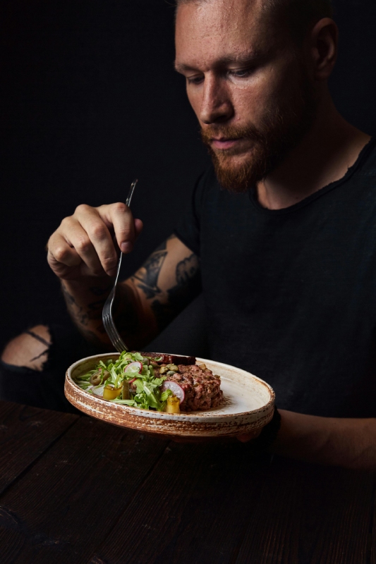 Man with a beard and tattoos holding a fork, about to eat a gourmet dish on a rustic plate in a dark setting.