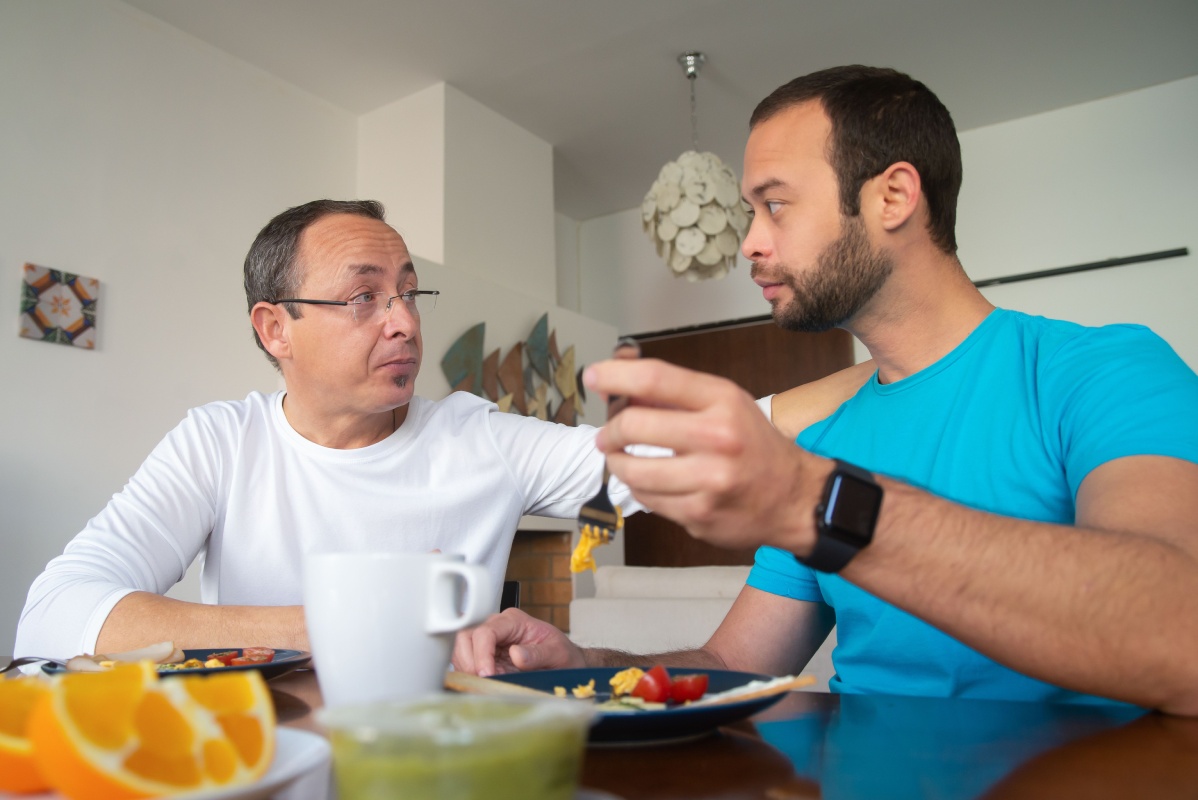 Two men eating breakfast together at a table with plates of food, a cup, and sliced oranges in a bright room.