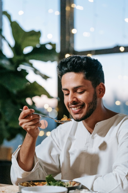 Man in white shirt eating food with a spoon in a softly lit room with plants and window lights in the background.