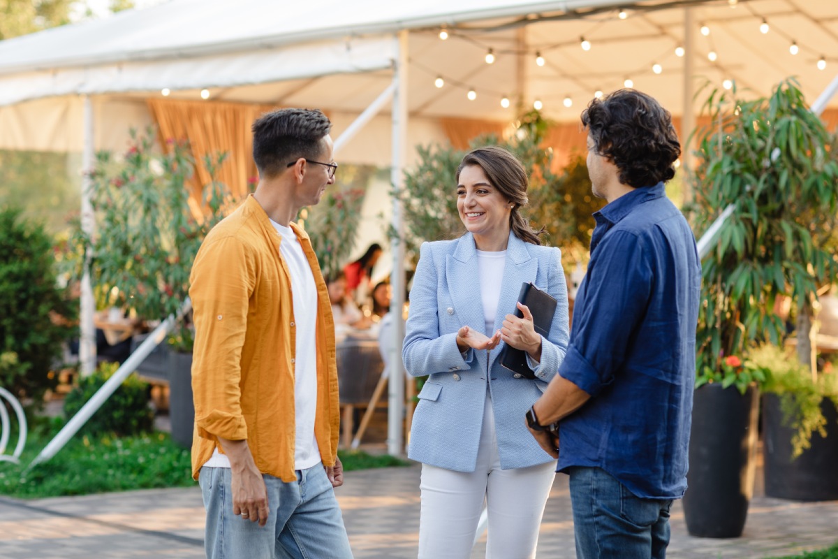 A group of three people happily converses outdoors near a tent, surrounded by greenery. The setting is casual and lively, suggesting a social gathering.