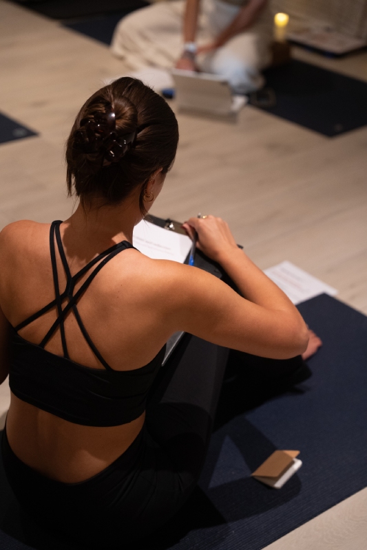 Woman in a black strappy workout top sits on a yoga mat, writing in a notebook. Another person is in the background, seated beside a candle, creating a calm atmosphere.