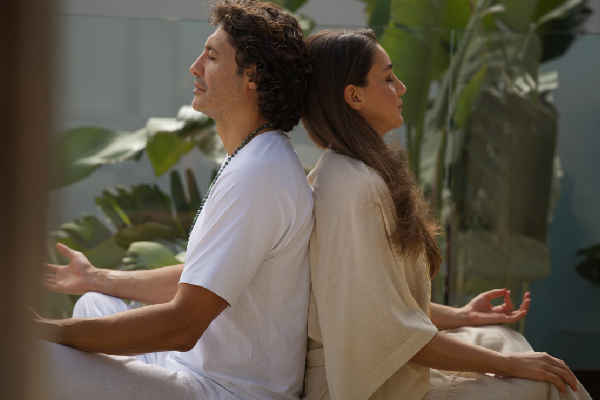 A man and woman sit back-to-back in meditation, eyes closed, in a peaceful garden. They're wearing white and beige, conveying calm and serenity.