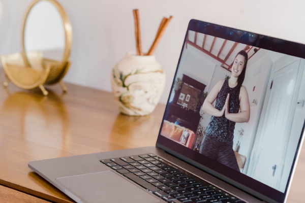 A laptop on a wooden table displays a cheerful person doing yoga, hands in a prayer pose. Nearby, a decorative vase and a basket add warmth to the setting.