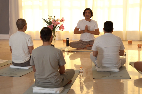 A group meditates on yoga mats in a serene room with soft lighting. An instructor sits cross-legged, leading the session with hands in prayer.