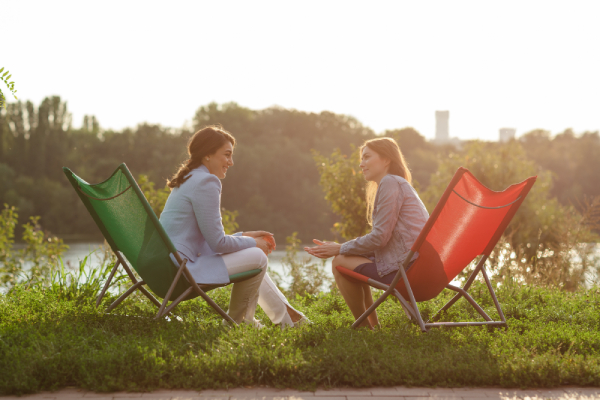 Two women sit in colorful deck chairs, engaged in conversation by a river, with a serene grassy landscape and trees under a warm, setting sun.