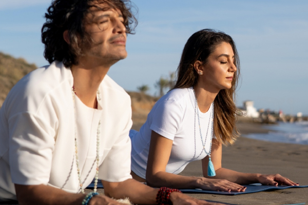Two people practice yoga on a tranquil beach. They are in a relaxed Cobra pose, eyes closed, wearing white tops and malas, embracing peace and mindfulness.