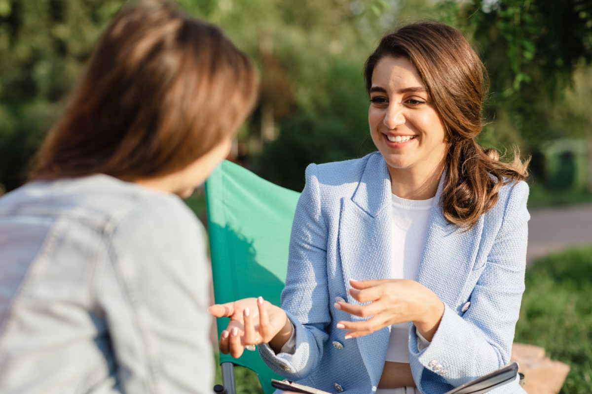 Two women sit outdoors having a friendly conversation. The woman in a blue blazer smiles warmly, gesturing with her hands. The atmosphere is relaxed and sunny.