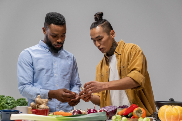 Two men are focused on preparing food in a kitchen. They are surrounded by fresh produce like kale, carrots, and a pumpkin on the counter.