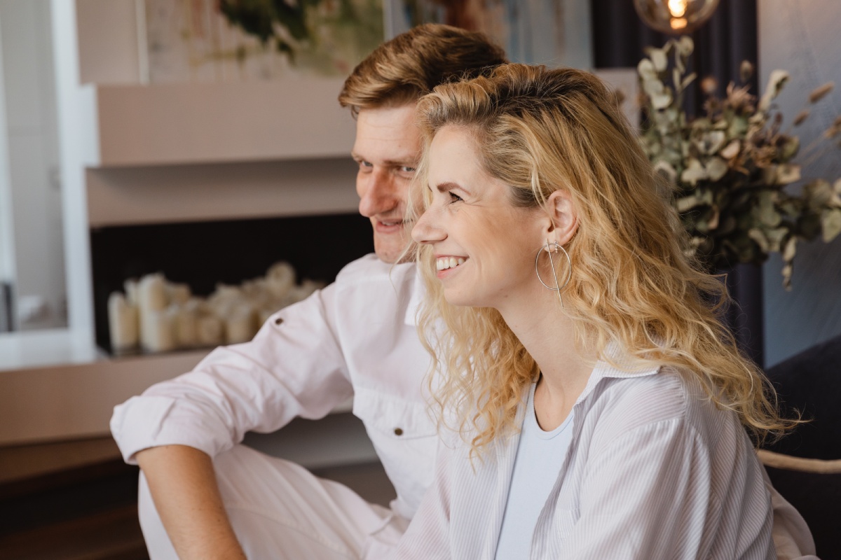 Couple sitting closely indoors, woman with curly blonde hair wearing hoop earrings, man dressed in white shirt and pants.