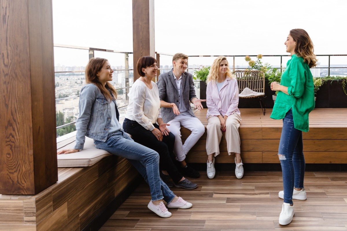 A group of five people are on a rooftop patio with a city view. Four are seated on a bench, engaged and smiling toward the standing woman, creating a lively and friendly atmosphere.